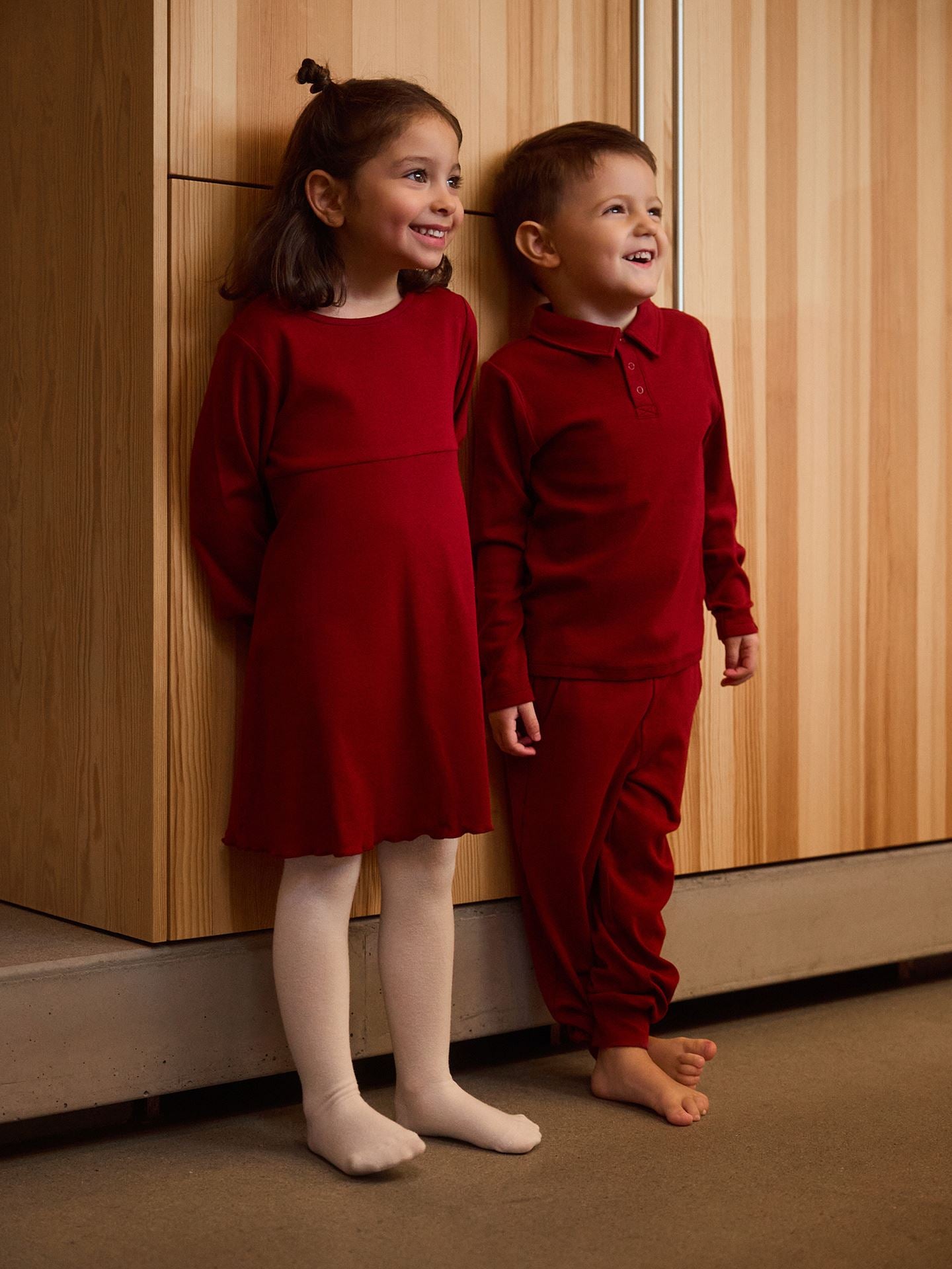Two children in matching red outfits standing against a wooden wall.