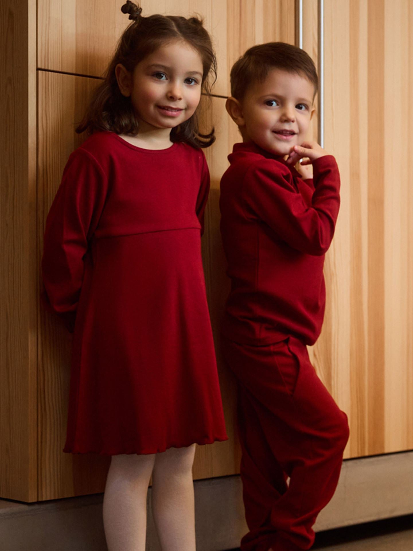 Two children in red outfits standing against a wooden wall.