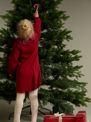 Child in a red dress reaching up to a Christmas tree, with a present at the base.